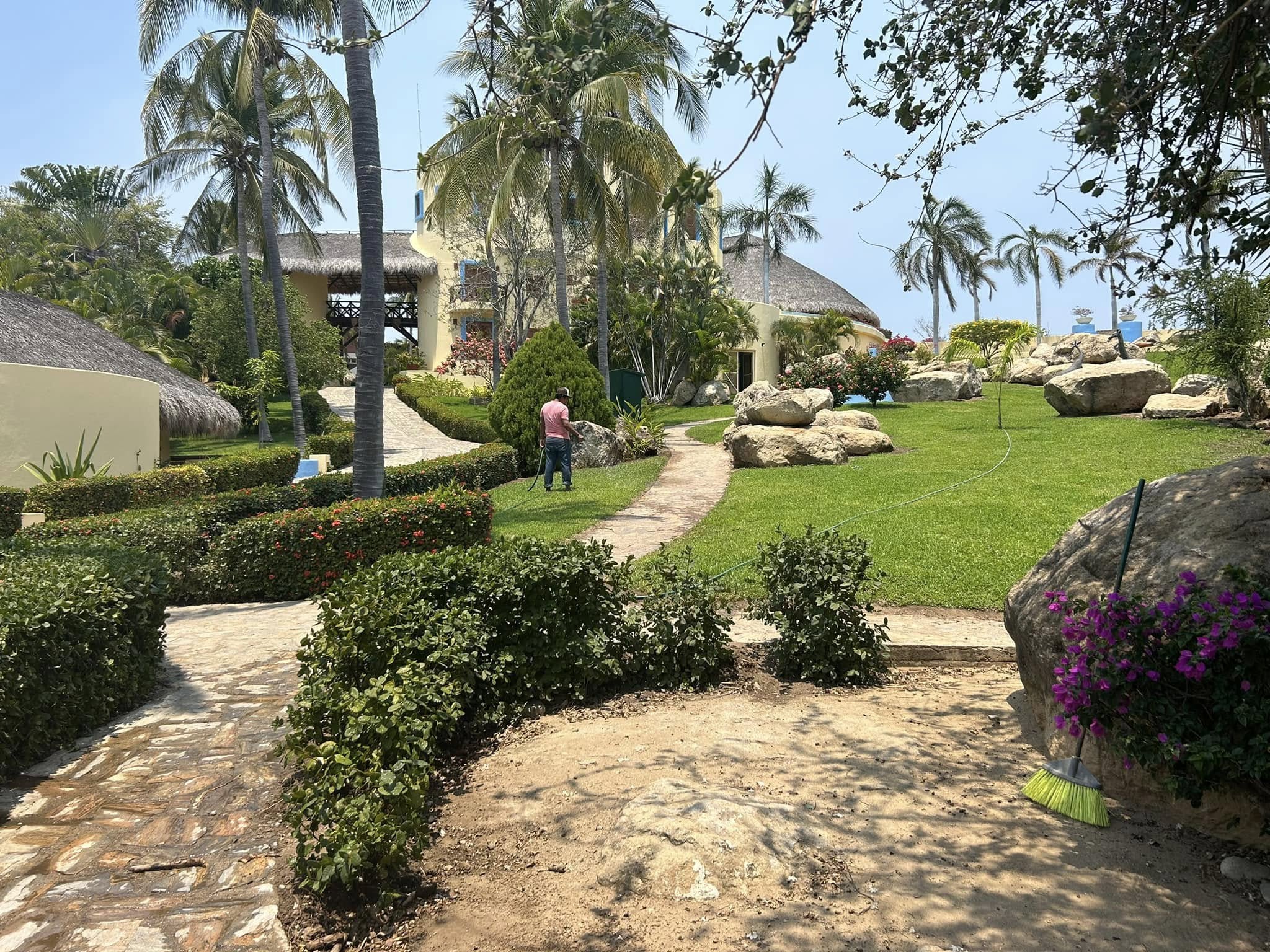 Jardín tropical frente a casa de playa con palmeras, rocas grandes y buganvilia, obra de El Edén